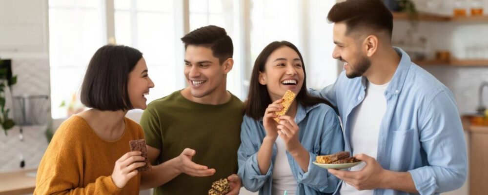 Group of friends laughing around a table filled with assorted protein snacks and smoothies